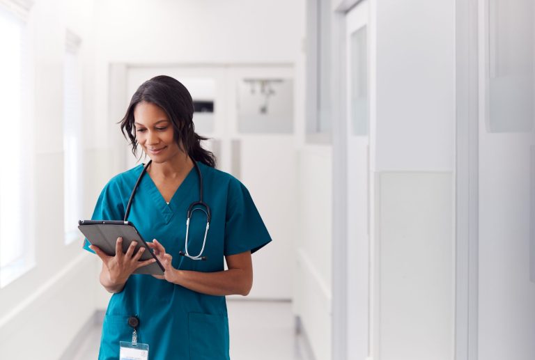 Portrait Of Smiling Female Doctor Wearing Scrubs In Hospital Corridor Holding Digital Tablet